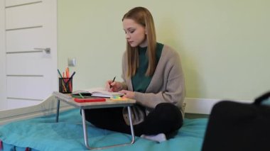 Genç bir öğrenci yatağında rahatça otururken derslerine odaklanır. Notlar yazıyor ve kırtasiyesini düzenliyor. Öğrenmek için mükemmel barışçıl bir ortam yaratıyor..