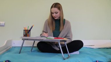 Genç bir kadın, renkli kalemlerle çizim becerilerine odaklanarak paspasın üzerinde rahatça oturur. Oda parlak ve davetkar, yaratıcılığını ve tutkusunu sergiliyor..