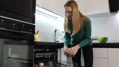 Modern bir mutfakta bir kadın yemek hazırladıktan sonra bulaşık makinesine temiz bulaşık koyar. Mekan düzenli ve mutfak malzemeleriyle dolu. Ev düzenine bağlılığını gösteriyor..