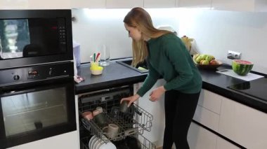 Parlak bir mutfakta, bir kadın üretken bir günün tadını çıkarırken bulaşık makinesini boşaltır, bardak ve tabakları düzenler. Yakınlardaki tezgahta taze meyve sergileniyor..