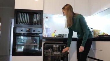 Bir kadın, çağdaş bir mutfakta bulaşık makinesine temiz bulaşıklar koyuyor. Doğal ışık boşluğu dolduruyor, güne hazırlanırken verimli sabah rutinini vurguluyor..