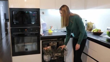 Bir kadın, çağdaş mutfağında bulaşık makinesinden bulaşık yıkar. Taze meyveler tezgahın üzerinde, o öğle yemeğini hazırlarken, parlak bir ev ortamında verimli bir çoklu görev sunuyor..