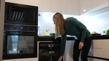 Bir kadın şık bir mutfakta yemek hazırlıyor, bulaşık makinesini kontrol ederek ve malzemeleri düzenleyerek birden fazla iş yapıyor. Güneş ışığı uzayı nazikçe aydınlatır ve sıcak bir atmosfer yaratır..