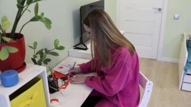 Genç bir kadın, düzenli çalışma alanındaki çeşitli renkli materyalleri kullanarak sanat projesine odaklanmış durumda. Etrafı bitki ve malzemelerle çevrili tahta bir masada rahatça oturuyor..