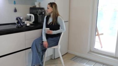 Bir kadın, kahvesini yudumlarken şık ve şık bir mutfakta sakin bir anın tadını çıkarır. Güneş pencereden içeri girerken rahat bir şekilde oturur ve güne huzurlu bir başlangıç yapar..