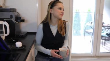 Genç bir kadın parlak bir mutfakta, elinde sıcak bir fincan kahveyle rahatça oturuyor. Büyük pencerelerden içeri güneş ışığı girerken gülümsüyor ve neşeli bir atmosfer yaratıyor..
