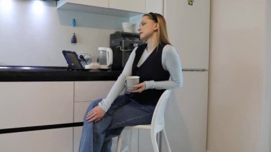 Bir insan şık bir mutfakta tabureye oturur, kahvesini yudumlar ve tablete bakar. Parlak ışıklandırma boşluğun nötr renklerini ve modern tasarımını vurgular.