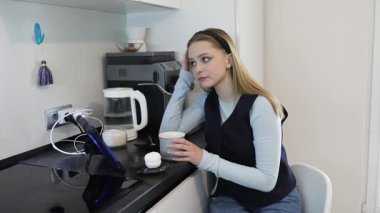 Genç bir kadın modern bir mutfakta oturur, derin düşüncelere dalmış bir fincandan çay yudumlar. Yumuşak atmosfer mutfak aletleri ve rahat bir titreşim ile tamamlanır..