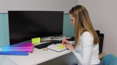 Bir kadın ofis masasında otururken derslerine odaklanır. Bir not defterine yazıyor ve renkli klasörler ve bilgisayarla çevrili yapışkan notlar kullanıyor..