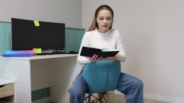 Kapalı bir ortamda, genç bir kadın mavi bir sandalyeye oturur, defterden notlar okur ve derslerine odaklanır. Atmosfer sakin ve düzenli, öğrenmek için mükemmel..