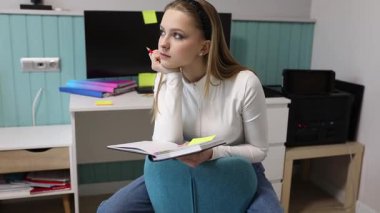 Genç bir kadın rahat bir odada oturur, bir defterden ders çalışırken çenesini eline koyar. Masasının üzerine renkli yapışkan notlar saçılmış, verimli bir atmosfer yaratıyor..