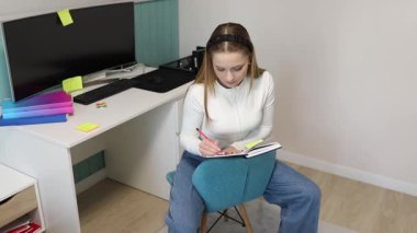 Mavi bir sandalyede oturan bir kız, renkli yapışkan notlarla çevrili ve dikkatle defterine odaklanmış, çalışma masasında çalışıyor..