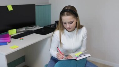 Genç bir kadın düzenli bir çalışma alanındaki bir deftere notlar yazarken derslerine odaklanıyor. Etrafı renkli yapışkan notlar ve bilgisayar ile çevrili. Üretken bir ortam yaratıyor..