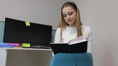 Genç bir kadın, modern bir odada dizüstü bilgisayarı ve renkli okul malzemeleriyle rahat rahat oturuyor. Çalışırken ışık konsantrasyonunu aydınlatıyor..