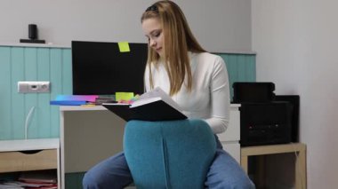 Genç bir kadın, rahat bir odada modern bir taburede oturup derslerine odaklanıyor. Minimalist dekor ve bilgisayarla çevrili bir deftere notlar yazıyor..