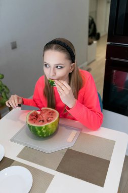 Saç bandı takan genç bir kız mutfak masasında oturup bir dilim karpuzun tadını çıkarıyor. Neşeli atmosfer ve canlı renkler bu ferahlatıcı yaz ikramının neşesini arttırır..