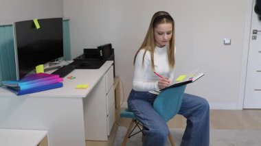 Odaklanmış genç bir kadın, renkli notlar, televizyon ve çalışma masası ile çevriliyken düzenli bir odada bir sandalyeye oturur ve bir deftere yazı yazar..