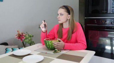 Genç bir kız, mutfak masasındaki oyulmuş bir karpuzdan yiyerek eşsiz bir yemek deneyiminden zevk alır. Parlak atmosfer eğlenceli aktiviteleri arttırır..