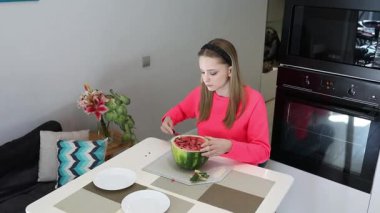 Parlak kazaklı genç bir kadın mutfak masasında karpuz kesiyor. Modern, rahat bir yerde sağlıklı bir atıştırmalığın tadını çıkarmaya hazırlanıyor..