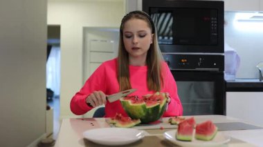 Modern bir mutfakta, neon tepeli genç bir kız ustalıkla bir karpuzu dilimler. Parçaları dikkatlice kaldırır ve sıcak bir yaz gününde sulu meyvenin tadını çıkarmaya hazırlanır..