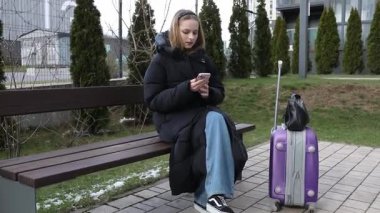 Genç bir kadın otobüs durağındaki bankta rahat bir şekilde oturuyor, üzerinde kalın bir ceket ve kot pantolon var. Çağdaş şehir atmosferinde yanında mor bir bavul dururken akıllı telefonunu kontrol ediyor..