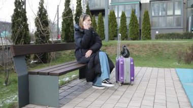 Parktaki bankta oturan bir kadın, sıcacık giyinmiş, telefonla konuşuyor. Renkli bavulu, birisini beklerken yerde yatar..