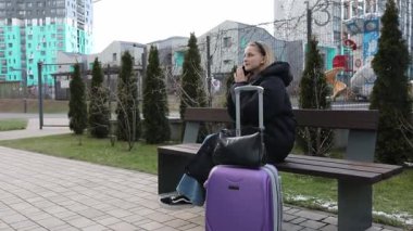 Şehir parkında bir bankta oturan genç bir kadın bekliyor. Yanında mor bir bavulu var ve telefonunu kullanıyor. Baharın başında modern binalar ve yeşilliklerle çevrili..