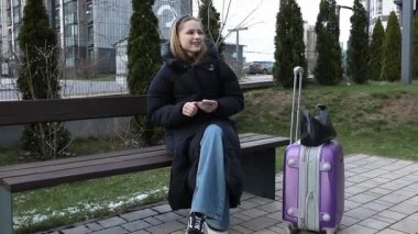 Genç bir kadın otobüs durağındaki bankta oturmuş, mor bavulu yanında dururken telefonunu kontrol ediyor. Çevrede binalar ve yeşillikler var..