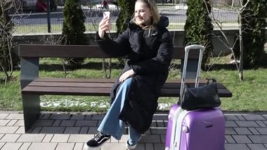 Genç bir gezgin parkta bir bankta oturuyor, gülümsüyor ve telefonuyla selfie çekiyor. Yanında mor bir bavulu var ve günlük kıyafetleriyle güneşli havanın tadını çıkarıyor..