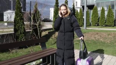 Uzun paltolu genç bir kadın şehir parkında bir bankın yanında duruyor, telefonuyla konuşuyor. Bir bavulu var ve güneşli havanın tadını çıkarırken sabırla bekliyor..