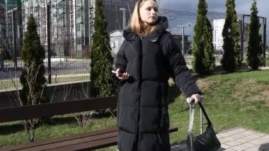 Siyah ceket giyen genç bir kadın, elinde akıllı bir telefon ve bir çanta tutarken güvenli bir şekilde kentsel alanda duruyor. Yeşilliklerle çevrili hoş havanın tadını çıkarıyor..