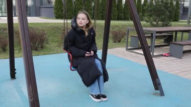 Rahat montlu genç bir kız, serin bir günde banliyöde sakin bir oyun alanıyla çevriliyken telefonuna odaklanarak salıncakta oturuyor..