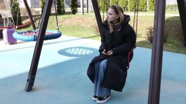 Samimi giyinmiş bir kız neredeyse boş bir oyun bahçesinde salıncakta sessizce oturuyor. Etrafı yeşilliklerle çevriliyken telefonunu tutarken düşünceli görünüyor..