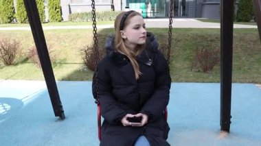 Genç bir kız parkta salıncakta oturuyor, üzerinde kalın bir palto ve elinde telefon var. Güneş parlıyor, yeşil çimenleri ve yolları aydınlatıyor..