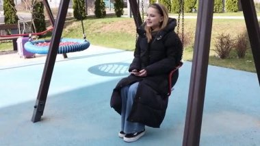 Genç bir kadın güneşli bir günde renkli bir parkta salıncakta rahatlıyor. Sıkı giyinmiş ve telefonuyla meşgul görünüyor. Dışarıda vakit geçiriyor..