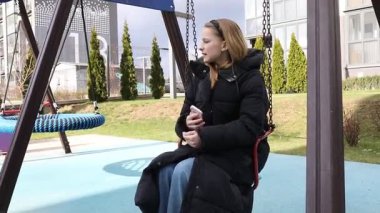 Genç bir kız, günümüzün çocuk parkında telefonuyla meşgul olarak salıncakta sallanmaktan hoşlanıyor. Kurulumda yemyeşil ve şık mimari yer alıyor..