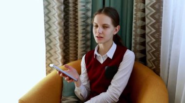 Genç bir kız akıllı telefonunu kullanırken sıcak, davetkar bir sandalyede oturuyor. Kapalı alan desenli perdeler ve rahat bir atmosfer yaratan yumuşak, renkli bir ortam içeriyor..