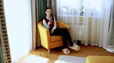 Genç bir birey, gündelik giyinmiş ve akıllı telefonuyla meşgul olan parlak, modern bir odada rahatça oturur. Uzay yumuşak ışıklandırma ve modern dekor içeriyor, sıcak bir atmosfer yaratıyor..