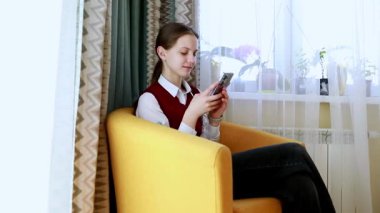 Genç bir kadın aydınlık bir odada rahat bir şekilde oturur ve akıllı telefonuna odaklanır. Kendi dünyasına dalmış, rahatlık ve memnuniyet yayıyor..