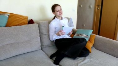 Bir kadın koltuğunda rahatça oturur ve ara sıra pozisyonunu düzeltirken kitap okumaya odaklanır. Konforlu oturma odasında sıcak renkler ve rahat bir dekor var..