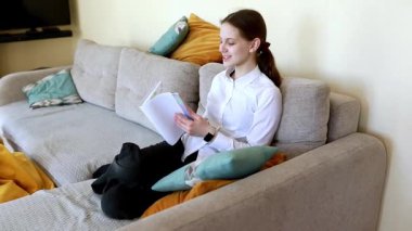 Düz saçlı genç bir kız rahat bir şekilde kanepede oturur, rahat bir oturma odasında renkli yastıklarla çevrili iken kitap okumakla meşgul olur. Sıcak atmosfer odaklanmasını sağlıyor..