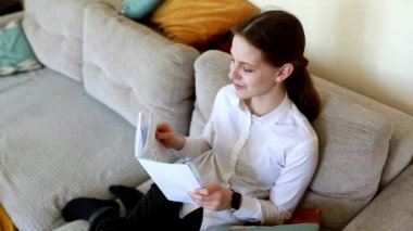 Genç bir kadın rahat bir şekilde kanepede oturur ve bir defter okur. Beyaz bir gömlek giymişti ve yumuşak yastıklarla çevrili, aktiviteleriyle meşgul görünüyordu..