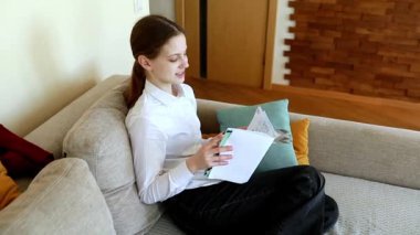 Genç bir kadın koltukta rahat bir şekilde oturuyor ve çeşitli belgeleri okumaya çalışıyor. Modern oturma odasında sıcak ahşap aksanlar ve rahat bir dekor var..