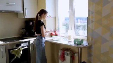 Genç bir kadın modern bir mutfağın penceresinde duruyor, sebze yıkıyor ve güneş ışığının tadını çıkarıyor. Alan temiz ve düzenli, dışarısı canlı görünüyor..
