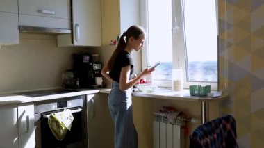 Genç bir kadın yemek hazırlarken modern bir mutfakta güneşli bir pencerenin yanında duruyor. Etrafı sebze ve çatal bıçaklarla çevriliyken telefonunu kontrol ediyor..