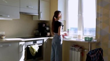 Genç bir kadın modern bir mutfakta, cam kenarındaki bir kasede malzemeleri karıştırıyor. Sabah ışığı mekanı dolduruyor, rahat ve davetkar bir atmosfer sergiliyor..