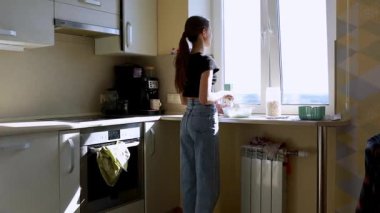 Günışığıyla dolu modern bir mutfakta, bir kadın yemek hazırlamakla, dışarıdaki manzaranın tadını çıkarmakla meşguldür. Hava sıcak ve davetkar..