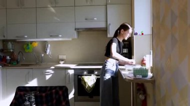 Parlak bir mutfakta, bir kız tezgahın önünde duruyor, yemek için malzemeleri dikkatlice hazırlıyor. Güneş ışığı içeri giriyor, odaklanmış tavrını ve etrafındaki organize alanı vurguluyor..