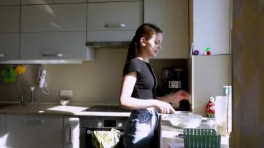 Genç bir kız iyi aydınlatılmış bir mutfakta yemek pişirirken bir kasede malzemeleri karıştırıyor. Büyük pencerelerden gelen güneş ışığı yemek pişirmek için sıcak ve davetkar bir atmosfer yaratıyor..