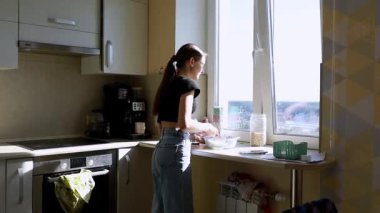 Genç bir kadın parlak bir mutfak tezgahında durup güneş ışığı pencereden içeri akarken malzemeleri hazırlıyor ve sıcak ve davetkar bir atmosfer yaratıyor..